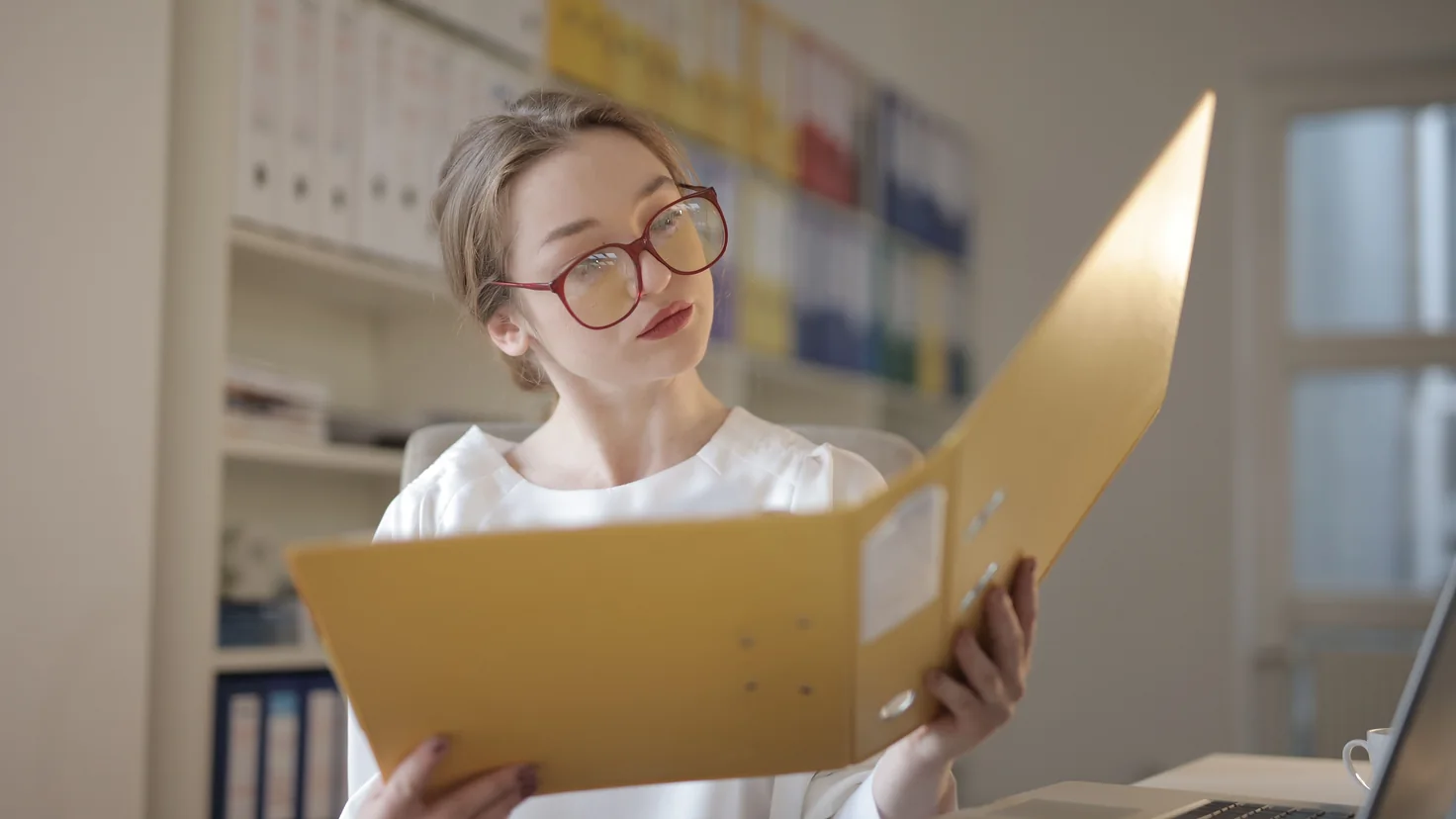 Sachbearbeiterin mit Brille blättert in einem Aktenordner, im Hintergrund ein Regal mit Ordnern